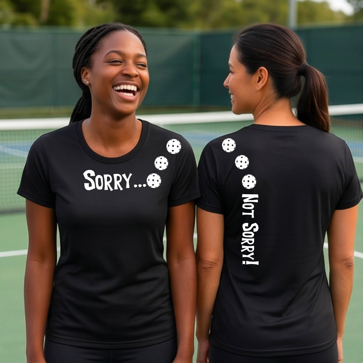 Two women on a pickleball court wearing black short-sleeve t-shirts with a front/back graphic design.  The front features the playful word “SORRY…” in bold white lettering, accented with three colorful pickleballs arranged diagonally across the upper chest.  The back design features “NOT SORRY!” in bold white lettering running vertically down the upper back, with three matching colorful pickleballs positioned above lettering.  