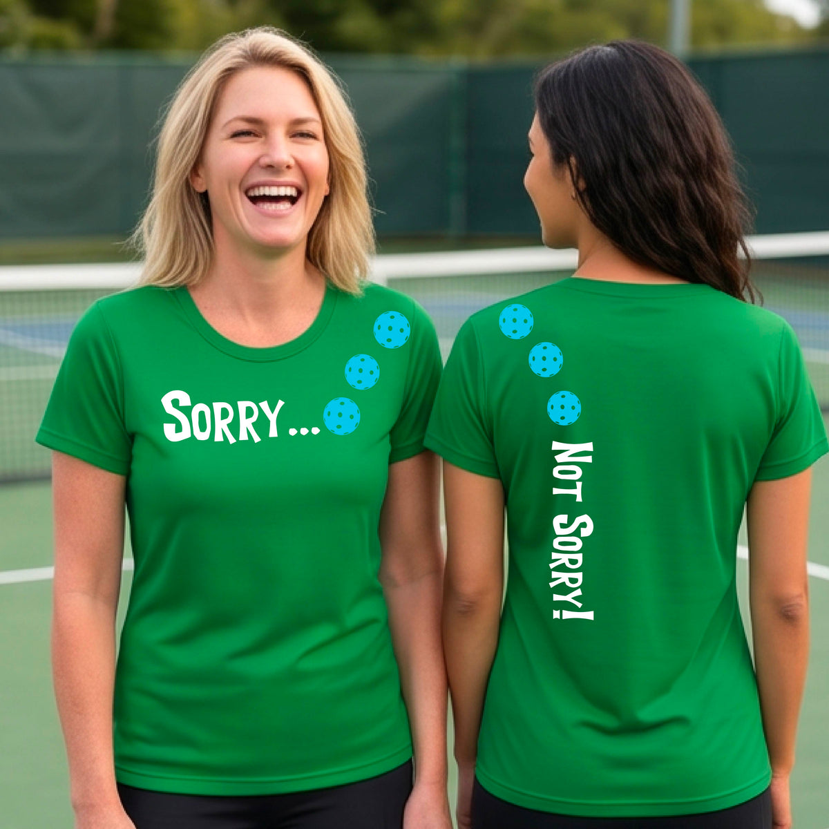 Two women on a pickleball court wearing green short-sleeve t-shirts with a front/back graphic design.  The front features the playful word “SORRY…” in bold white lettering, accented with three colorful pickleballs arranged diagonally across the upper chest.  The back design features “NOT SORRY!” in bold white lettering running vertically down the upper back, with three matching colorful pickleballs positioned above lettering.  