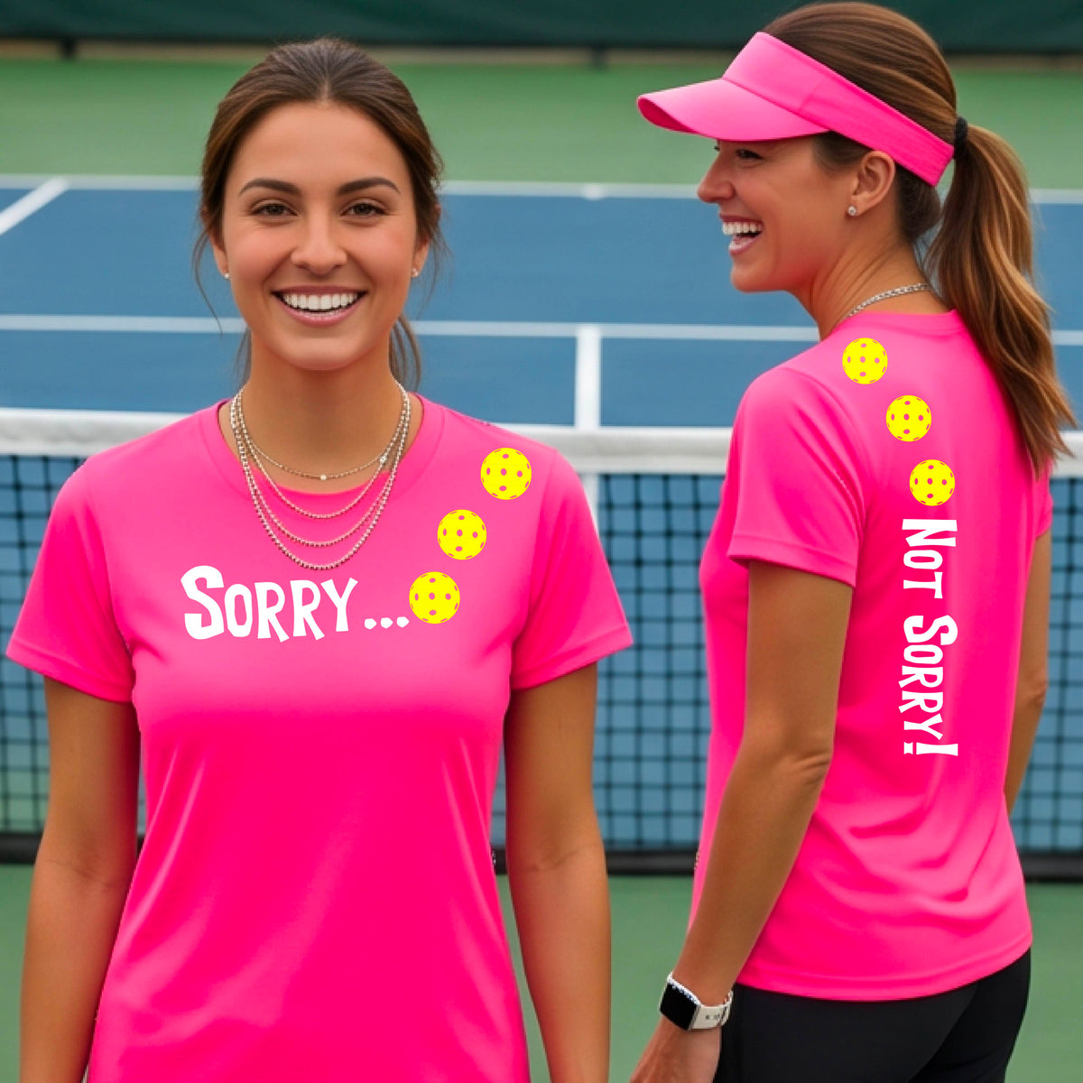 Two women on a pickleball court wearing pink short-sleeve t-shirts with a front/back graphic design.  The front features the playful word “SORRY…” in bold white lettering, accented with three colorful pickleballs arranged diagonally across the upper chest.  The back design features “NOT SORRY!” in bold white lettering running vertically down the upper back, with three matching colorful pickleballs positioned above lettering.  