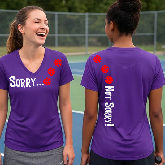 Two women on a pickleball court wearing purple short-sleeve v-neck t-shirts with a front/back graphic design.  The front features the playful word “SORRY…” in bold white lettering, accented with three colorful pickleballs arranged diagonally across the upper chest.  The back design features “NOT SORRY!” in bold white lettering running vertically down the upper back, with three matching colorful pickleballs positioned above lettering.  