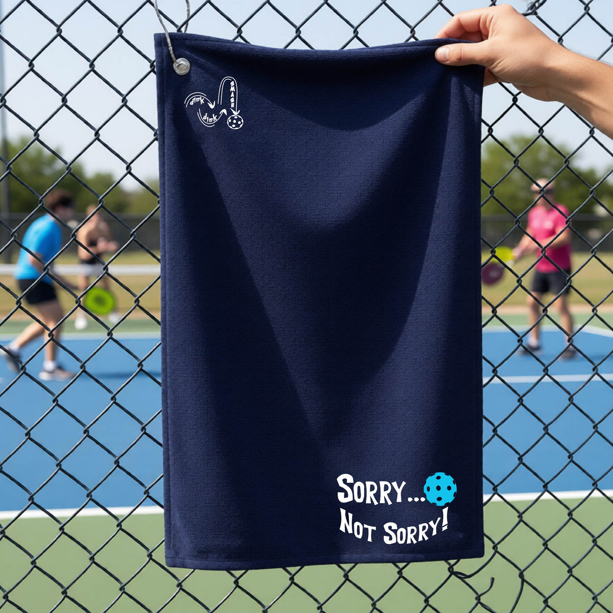 A close-up of a navy blue terry cloth grommet and hook towel hanging on a fence at a pickleball court, with pickleball motif design on the front and brim and features the playful word “SORRY…” in bold white lettering, accented a colorful pickleball, with the words 'Not sorry" curved underneath on the bottom right of the towel. .There is a white logo of 'DINK DINK SMASH' on the upper left of the towel.  