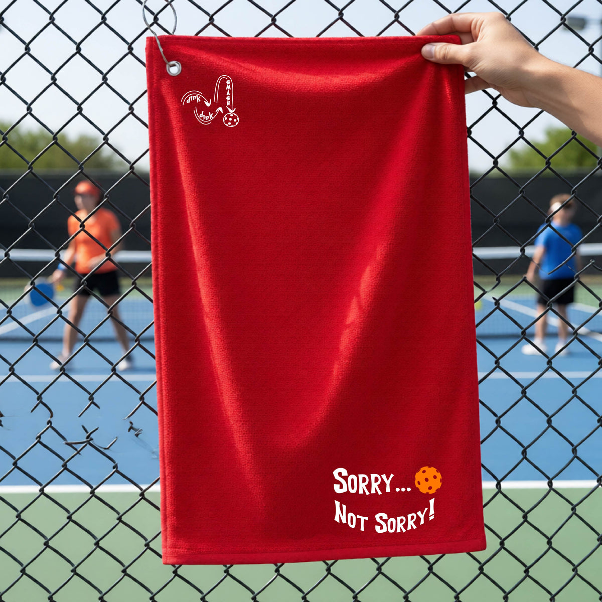 A close-up of a red terry cloth grommet and hook towel hanging on a fence at a pickleball court, with pickleball motif design on the front and brim and features the playful word “SORRY…” in bold white lettering, accented a colorful pickleball, with the words 'Not sorry" curved underneath on the bottom right of the towel. .There is a white logo of 'DINK DINK SMASH' on the upper left of the towel.  