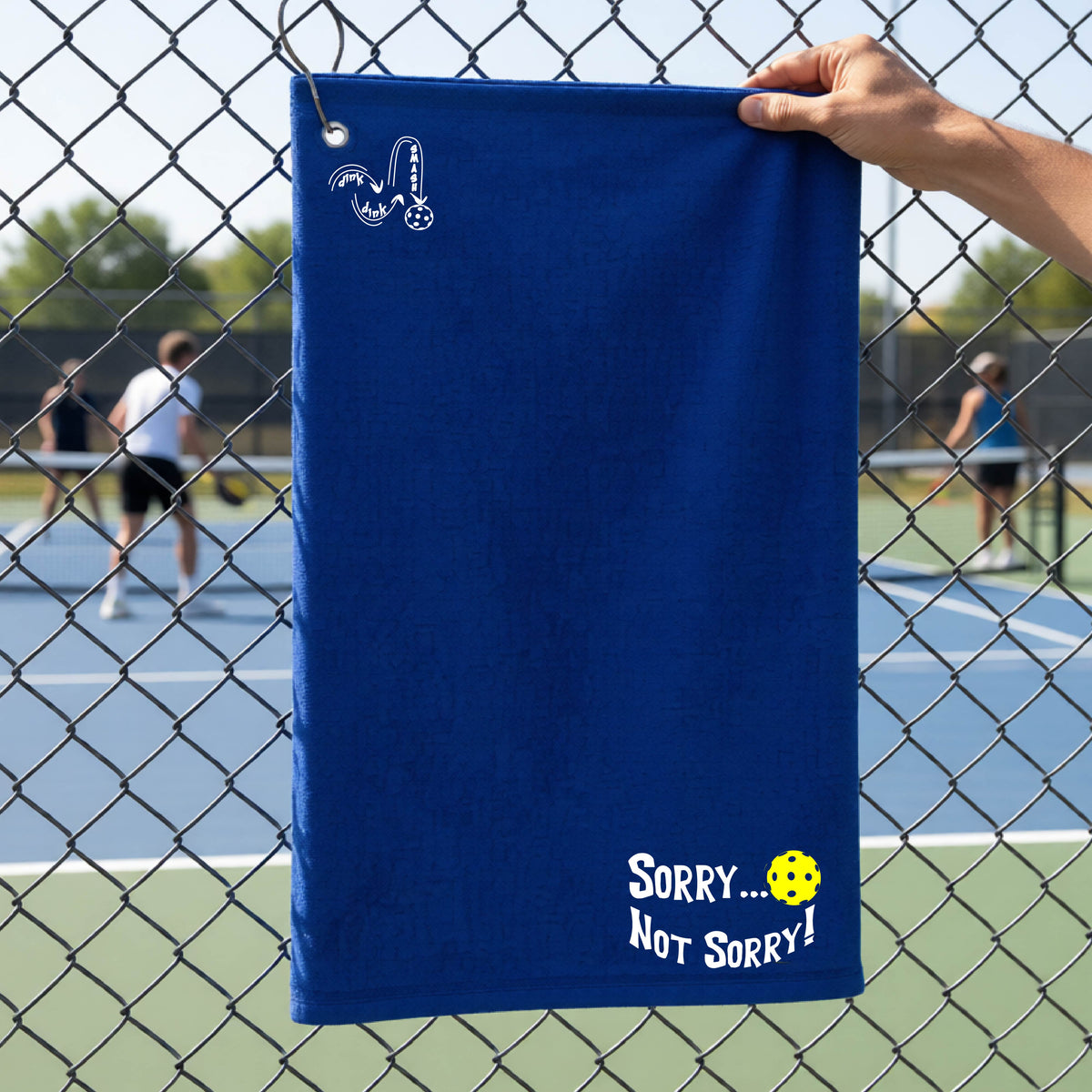 A close-up of a royal blue terry cloth grommet and hook towel hanging on a fence at a pickleball court, with pickleball motif design on the front and brim and features the playful word “SORRY…” in bold white lettering, accented a colorful pickleball, with the words 'Not sorry" curved underneath on the bottom right of the towel. .There is a white logo of 'DINK DINK SMASH' on the upper left of the towel.  