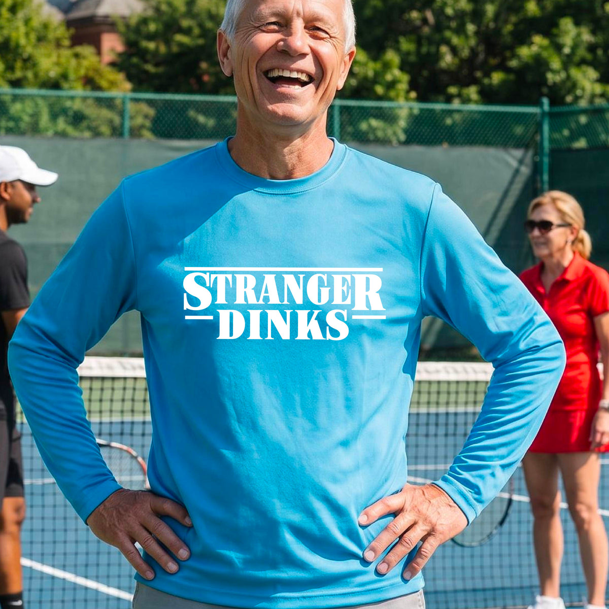 A man on a pickleball court wearing a bright blue long-sleeve t-shirt with a white graphic of the words “STRANGER DINKS” on the front of the shirt in the stule of the popular TV Series Stranger Things. 