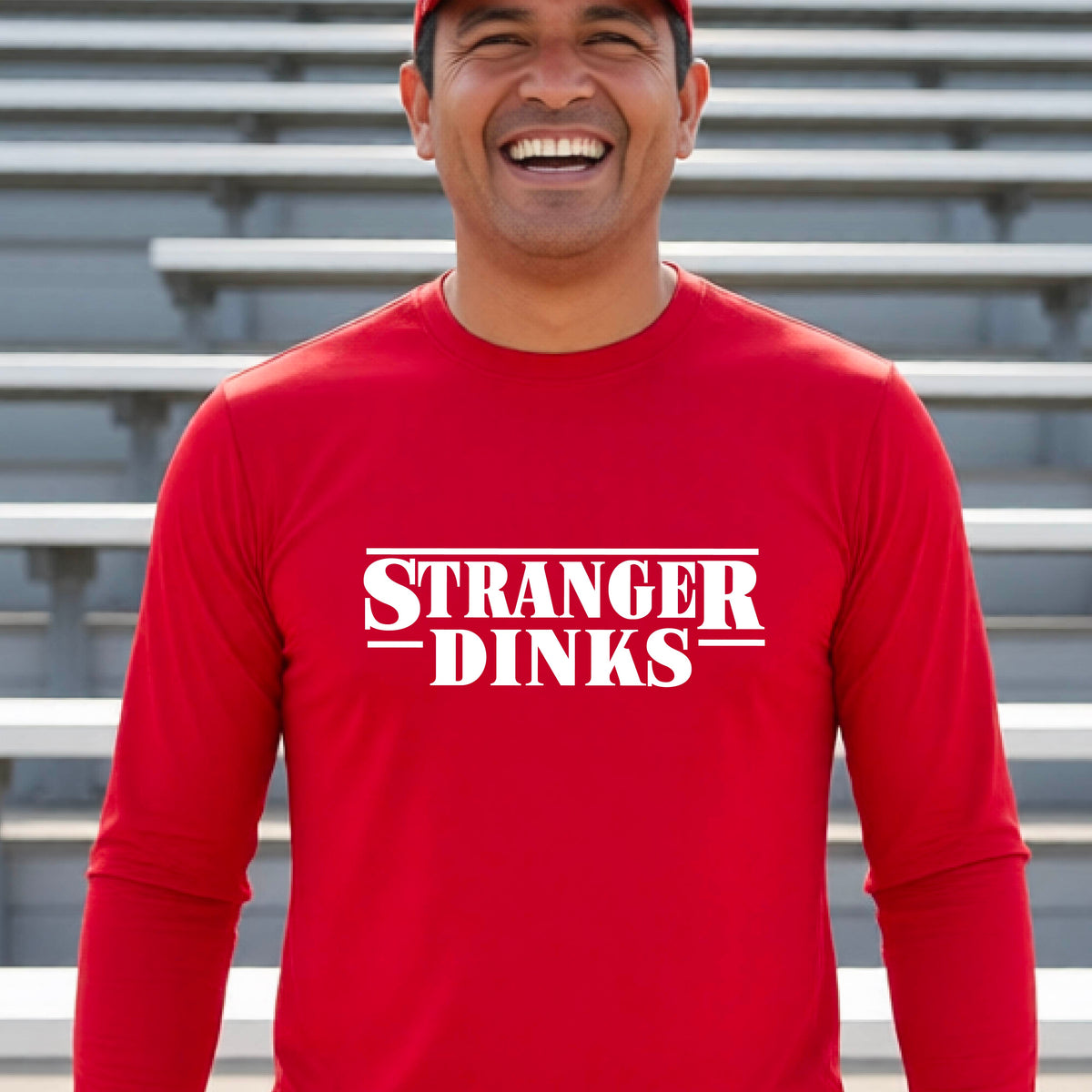 A man on a pickleball court wearing a red long-sleeve t-shirt with a white graphic of the words “STRANGER DINKS” on the front of the shirt in the stule of the popular TV Series Stranger Things. 