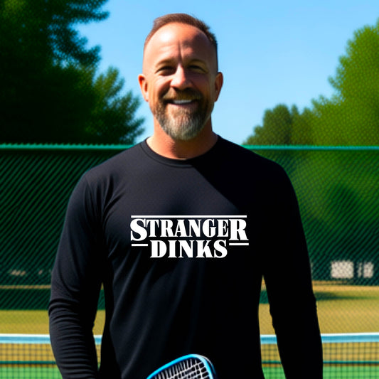 A man on a pickleball court wearing a black long-sleeve t-shirt with a white graphic of the words “STRANGER DINKS” on the front of the shirt in the stule of the popular TV Series Stranger Things. 

