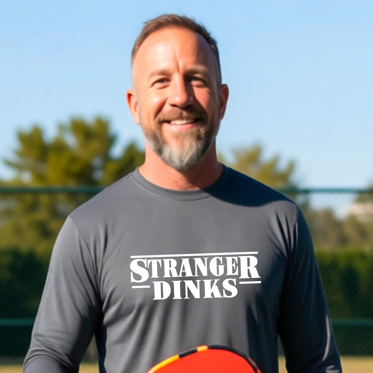 A man on a pickleball court wearing a gray long-sleeve t-shirt with a white graphic of the words “STRANGER DINKS” on the front of the shirt in the stule of the popular TV Series Stranger Things. 