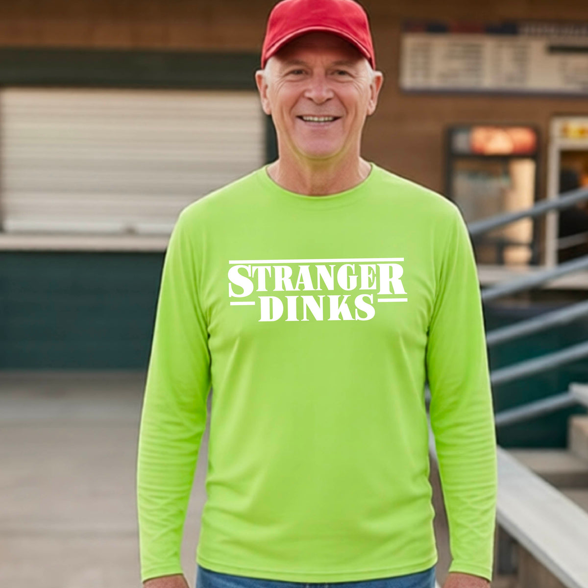 A man on a pickleball court wearing a lime green long-sleeve t-shirt with a white graphic of the words “STRANGER DINKS” on the front of the shirt in the stule of the popular TV Series Stranger Things. 