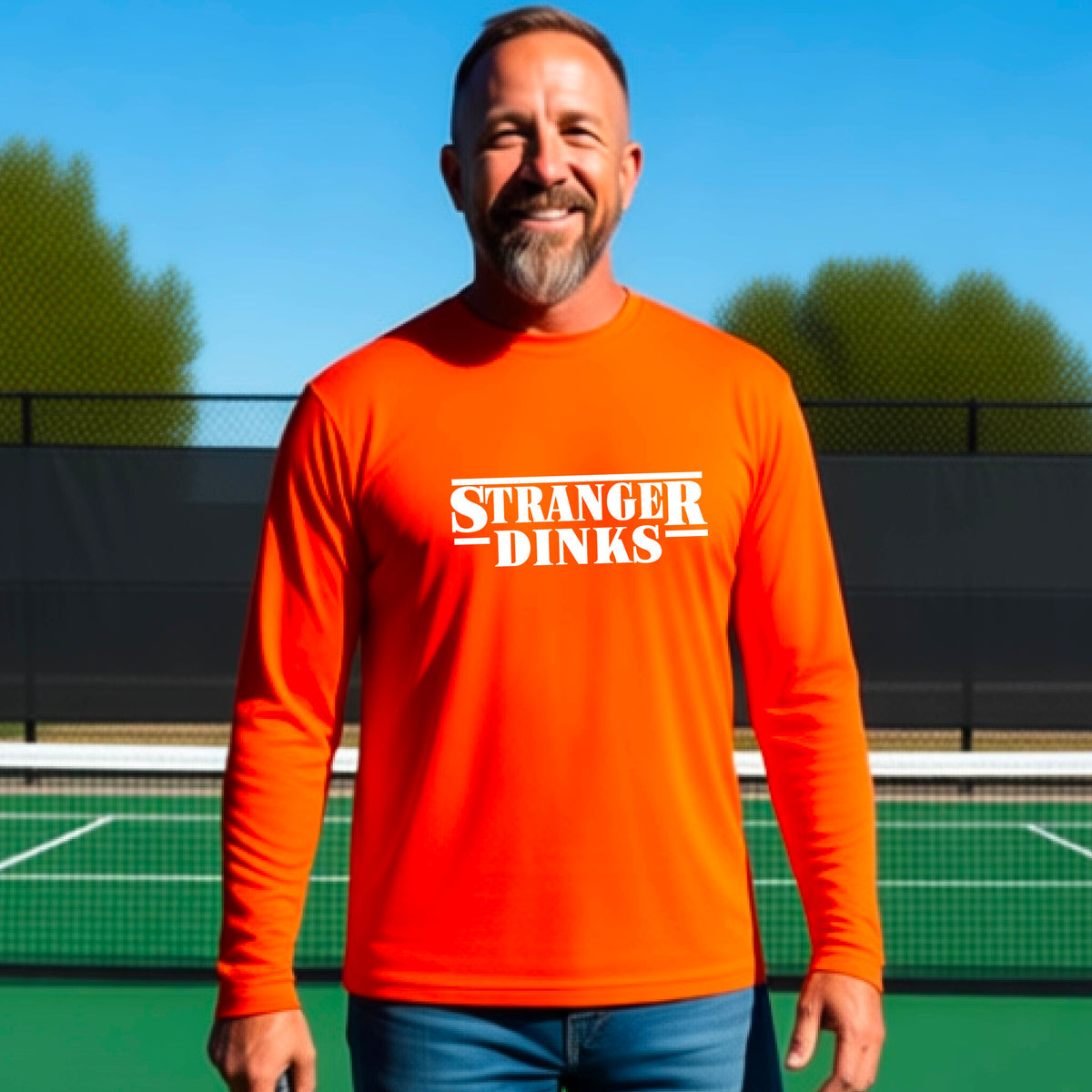 A man on a pickleball court wearing an orange long-sleeve t-shirt with a white graphic of the words “STRANGER DINKS” on the front of the shirt in the stule of the popular TV Series Stranger Things. 