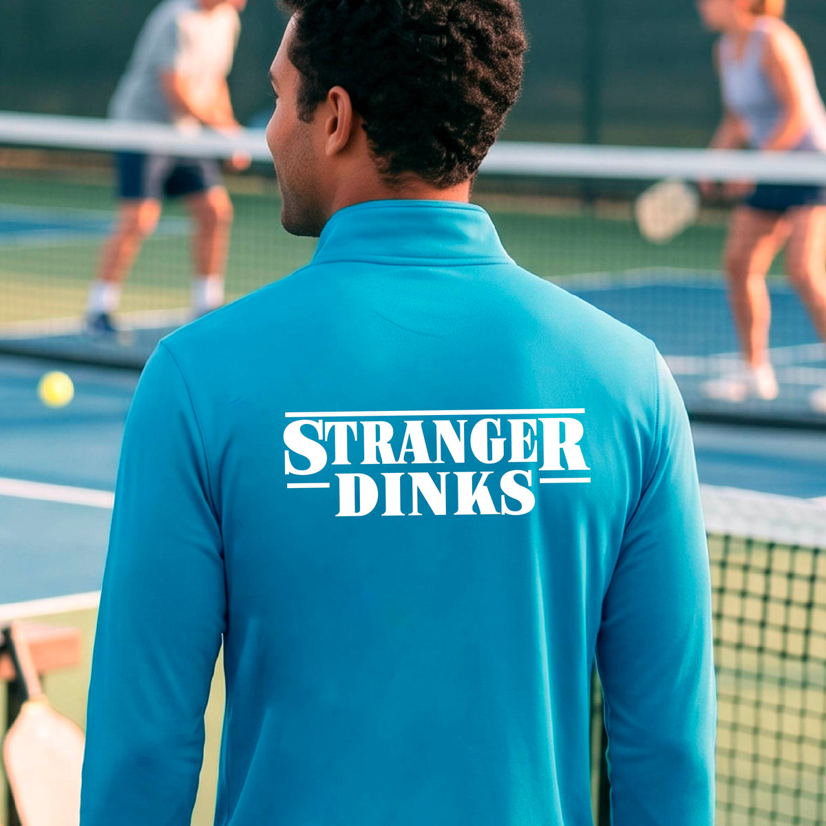 A man viewed from behind wearing a bright blue long-sleeve quarter-zip shirt shirt, standing in front of a pickleball court. The back of the shirt has a large white graphic that reads “STRANGER DINKS in the style of the TV series Stranger Things. 