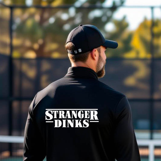 A man viewed from behind wearing a black long-sleeve quarter-zip shirt shirt, standing in front of a pickleball court. The back of the shirt has a large white graphic that reads “STRANGER DINKS in the style of the TV series Stranger Things. 