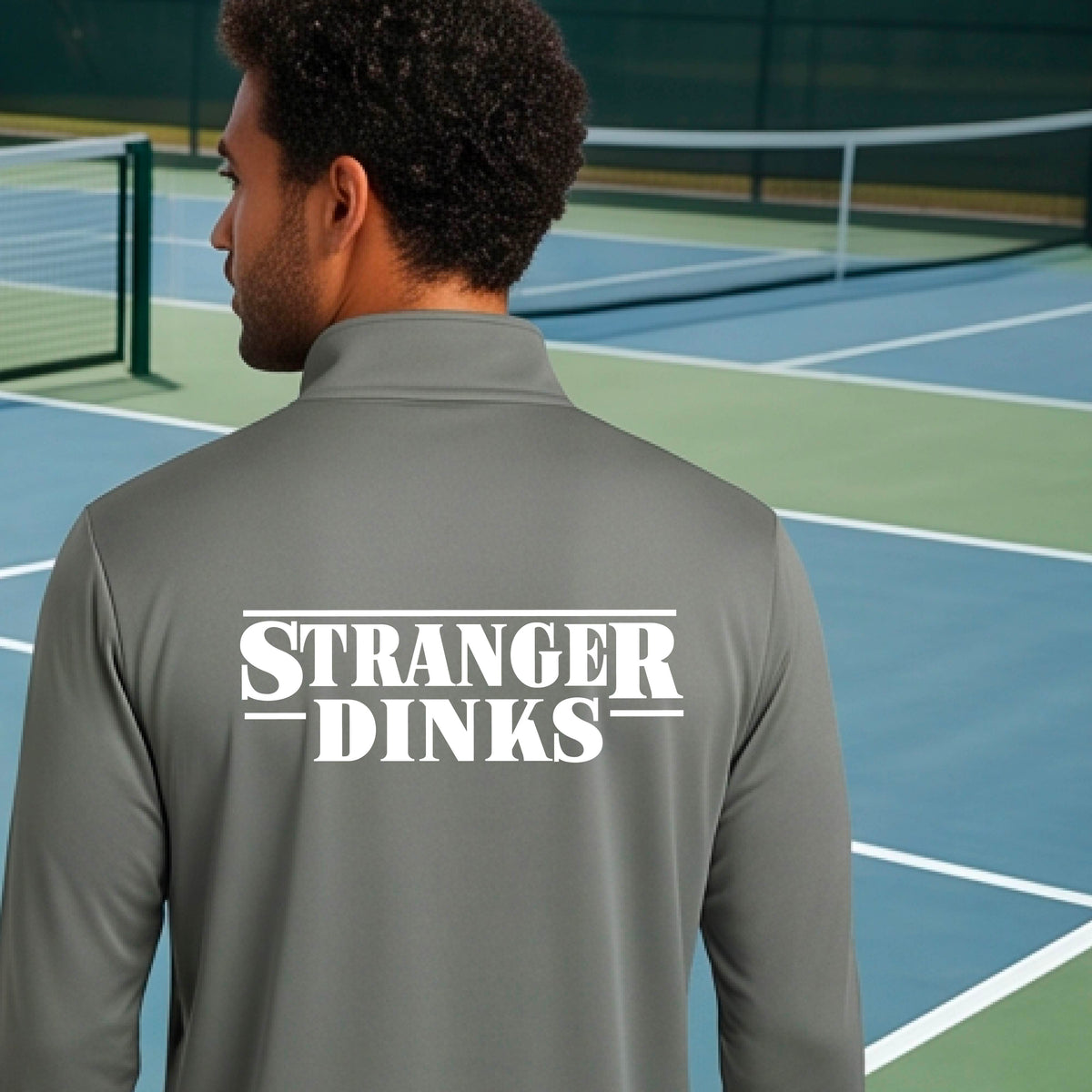 A man viewed from behind wearing a gray long-sleeve quarter-zip shirt shirt, standing in front of a pickleball court. The back of the shirt has a large white graphic that reads “STRANGER DINKS in the style of the TV series Stranger Things. 