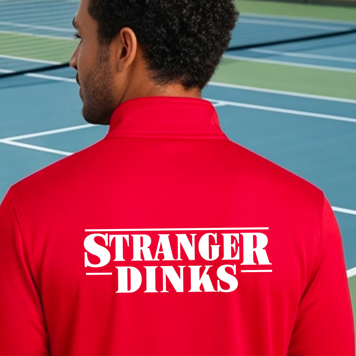 A man viewed from behind wearing a red long-sleeve quarter-zip shirt shirt, standing in front of a pickleball court. The back of the shirt has a large white graphic that reads “STRANGER DINKS in the style of the TV series Stranger Things. 