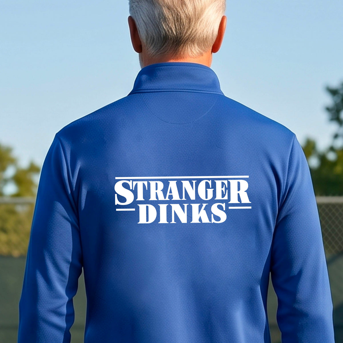 A man viewed from behind wearing a royal blue long-sleeve quarter-zip shirt shirt, standing in front of a pickleball court. The back of the shirt has a large white graphic that reads “STRANGER DINKS in the style of the TV series Stranger Things. 