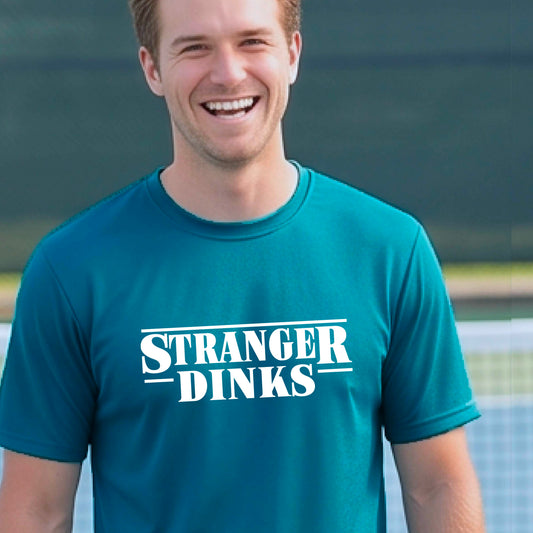 A man on a pickleball court wearing a teal short-sleeve t-shirt with a white graphic of the words  “STRANGER DINKS” on the front of the shirt in the stule of the popular TV Series Stranger Things. 