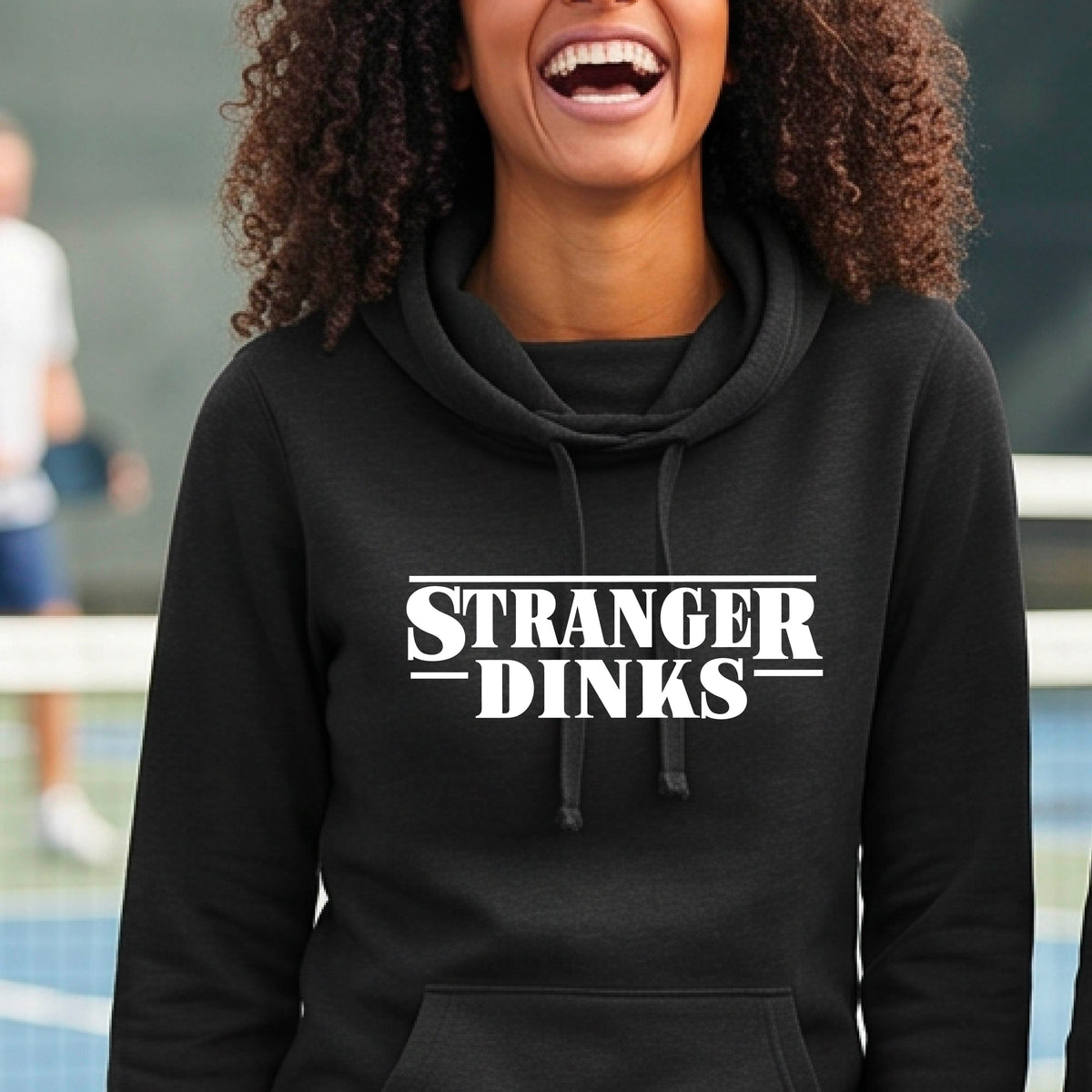 A woman wearing a black cowl-neck hoodie pullover with a white graphic of the words “STRANGER DINKS” on the front of the shirt in the style of the popular TV Series Stranger Things. 