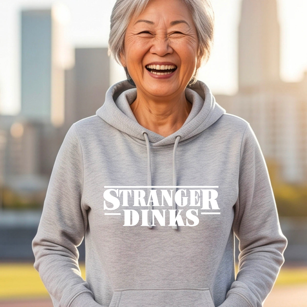 A woman wearing a light gray cowl-neck hoodie pullover with a white graphic of the words “STRANGER DINKS” on the front of the shirt in the style of the popular TV Series Stranger Things. 