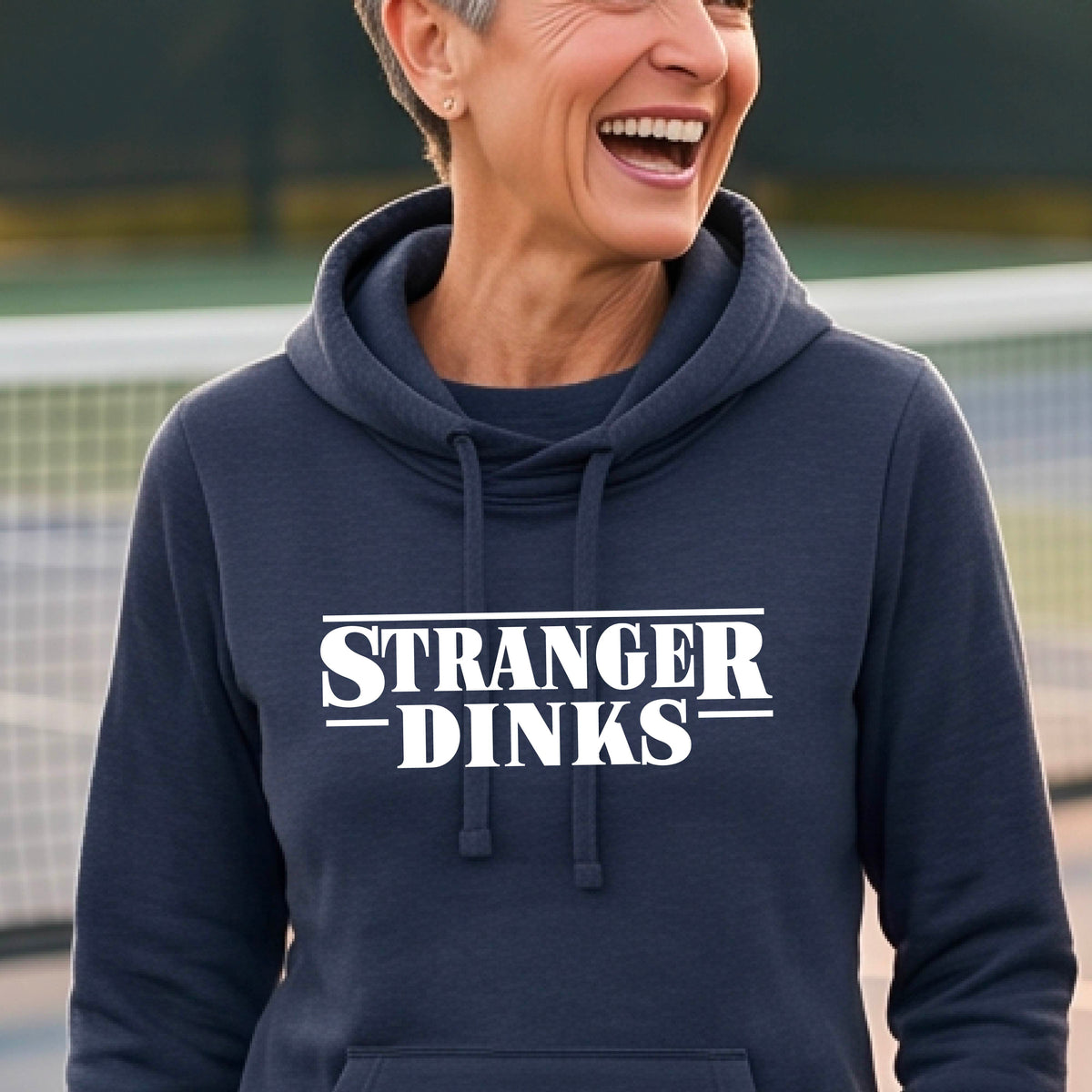 A woman wearing a navy blue cowl-neck hoodie pullover with a white graphic of the words “STRANGER DINKS” on the front of the shirt in the style of the popular TV Series Stranger Things. 