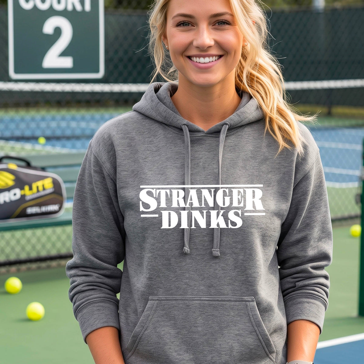 A woman on a pickleball court wearing a gray fitted hoodie pullover with a white graphic of the words “STRANGER DINKS” on the front of the shirt in the style of the popular TV Series Stranger Things. 