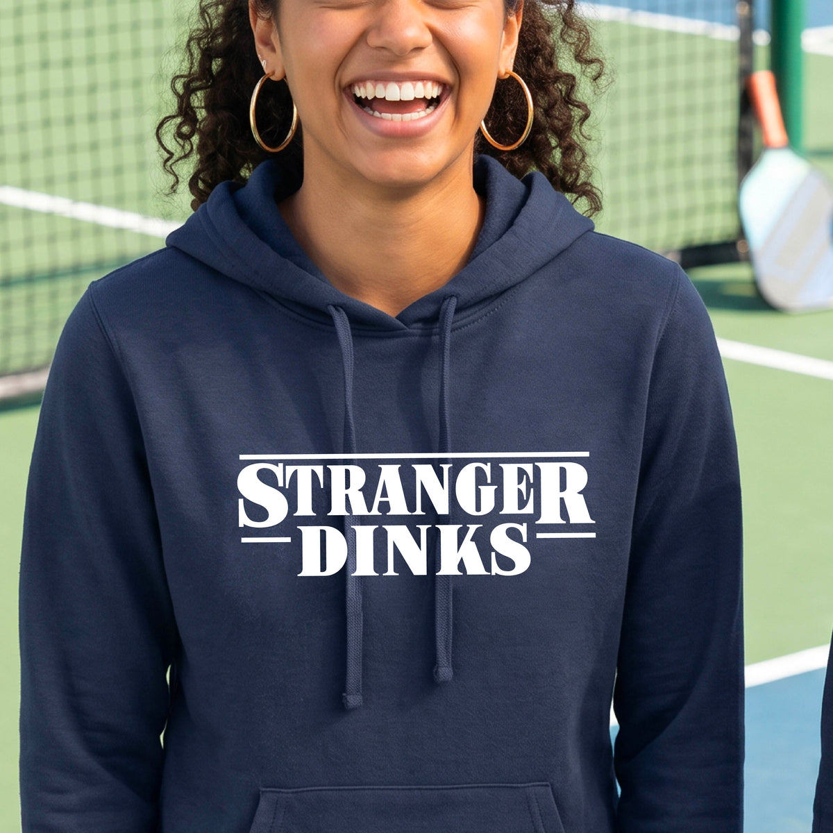 A woman on a pickleball court wearing a navy blue fitted hoodie pullover with a white graphic of the words “STRANGER DINKS” on the front of the shirt in the style of the popular TV Series Stranger Things. 