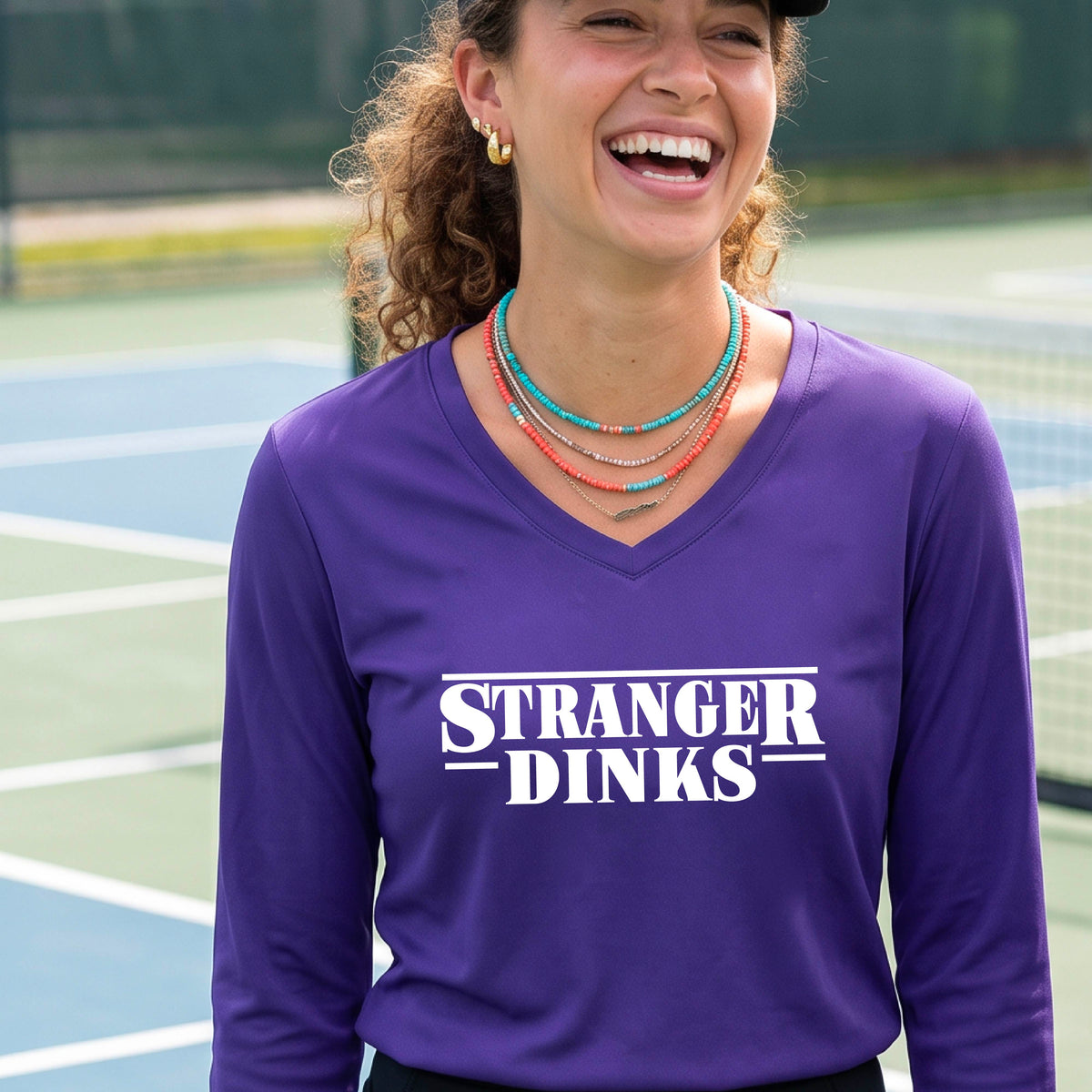A man on a pickleball court wearing a purple long-sleeve t-shirt with a white graphic of the words “STRANGER DINKS” on the front of the shirt in the style of the popular TV Series Stranger Things. 