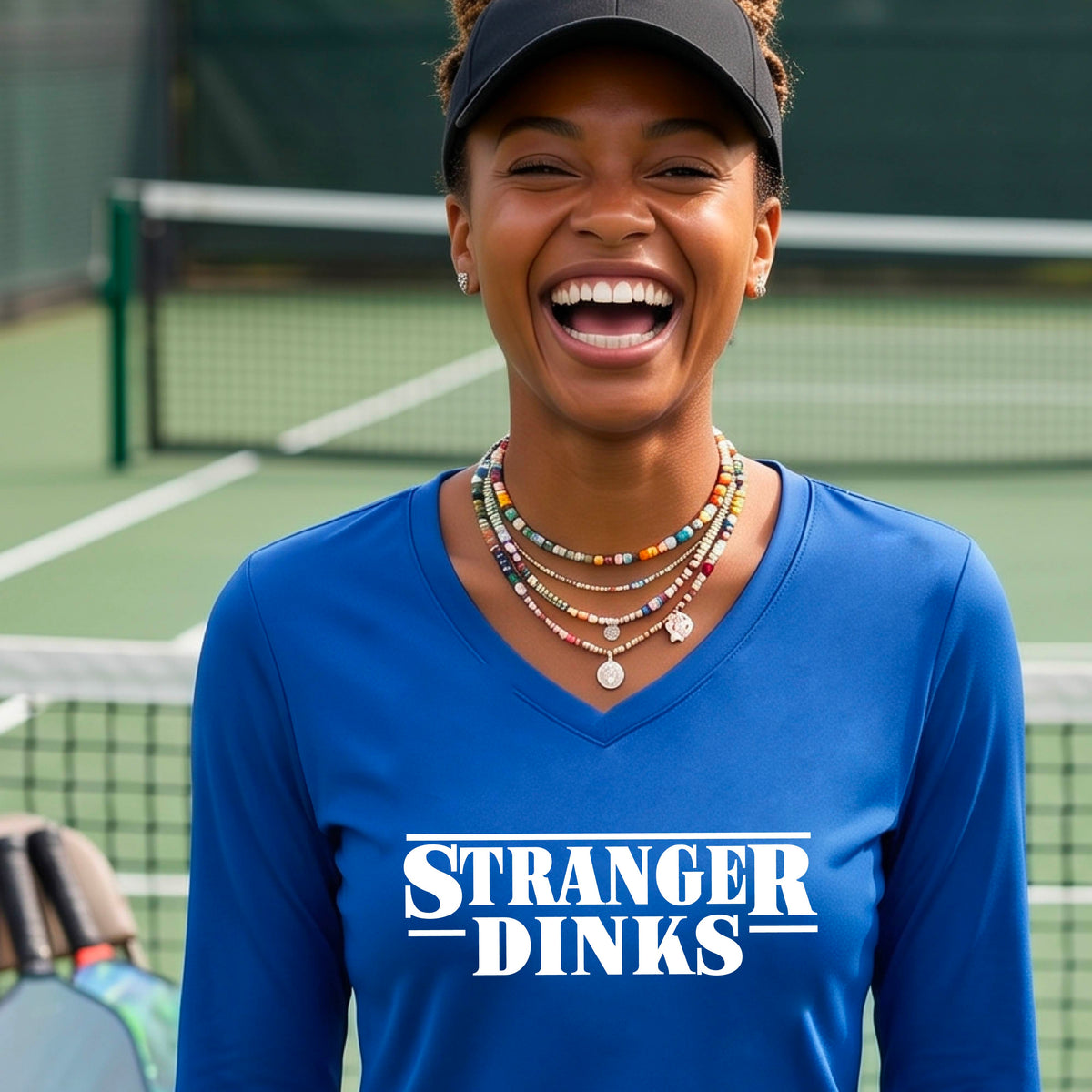 A man on a pickleball court wearing a royal blue long-sleeve t-shirt with a white graphic of the words “STRANGER DINKS” on the front of the shirt in the style of the popular TV Series Stranger Things. 