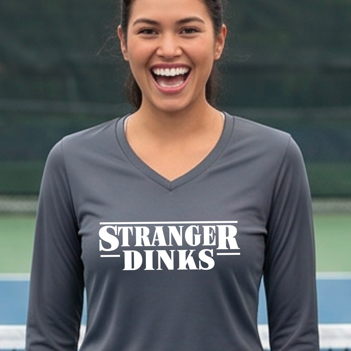 A man on a pickleball court wearing a dark gray long-sleeve t-shirt with a white graphic of the words “STRANGER DINKS” on the front of the shirt in the style of the popular TV Series Stranger Things. 