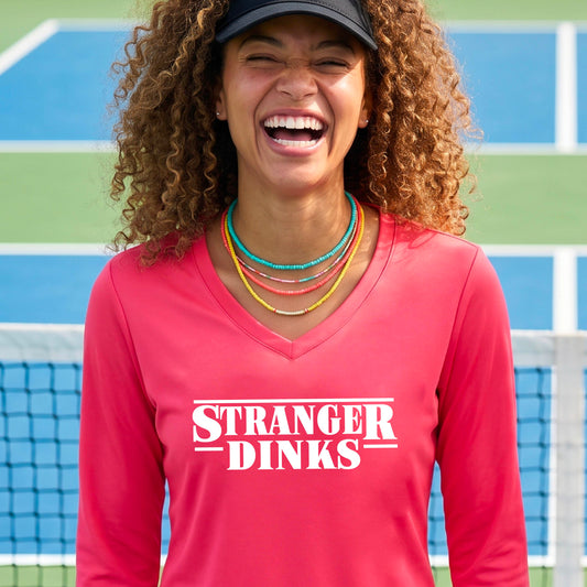A man on a pickleball court wearing a coral pink long-sleeve t-shirt with a white graphic of the words “STRANGER DINKS” on the front of the shirt in the style of the popular TV Series Stranger Things. 