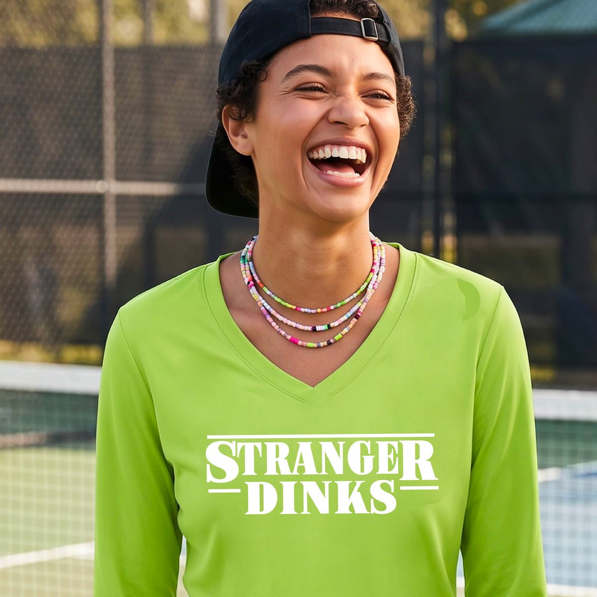 A man on a pickleball court wearing a lime green long-sleeve t-shirt with a white graphic of the words “STRANGER DINKS” on the front of the shirt in the style of the popular TV Series Stranger Things. 