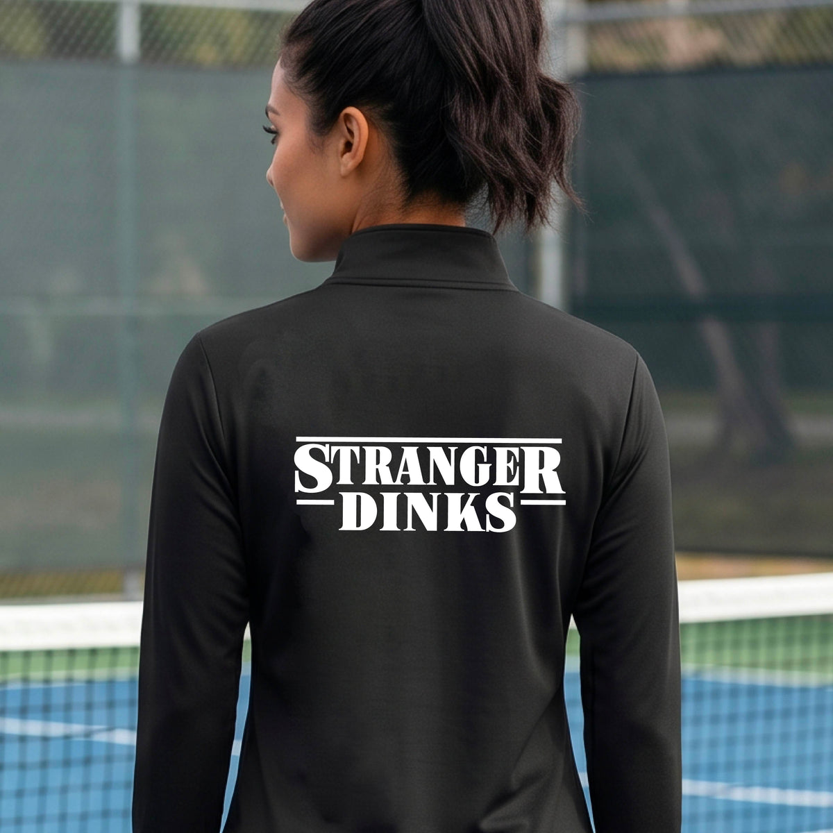 A person with short silver hair seen from behind wearing a black quarter-zip long-sleeve shirt with a white graphic of the words “STRANGER DINKS” on the front of the shirt in the style of the popular TV Series Stranger Things. 