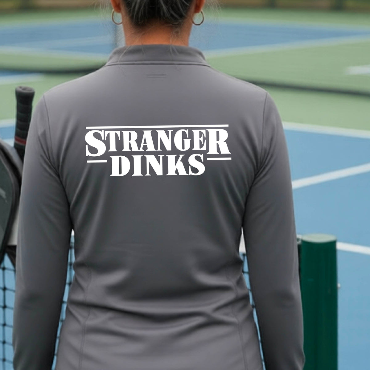 A person with short silver hair seen from behind wearing a gray quarter-zip long-sleeve shirt with a white graphic of the words “STRANGER DINKS” on the front of the shirt in the style of the popular TV Series Stranger Things. 
