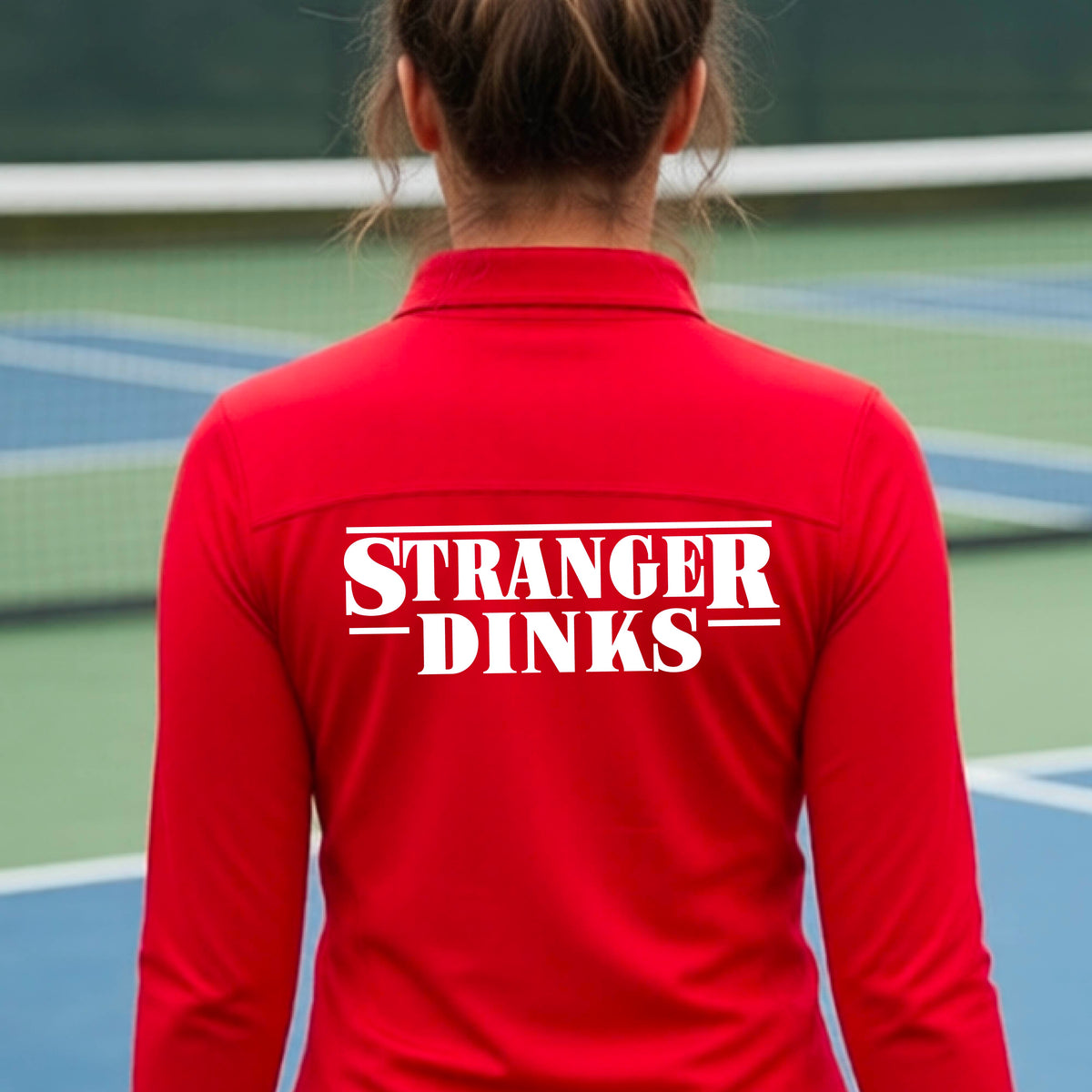 A person with short silver hair seen from behind wearing a red quarter-zip long-sleeve shirt with a white graphic of the words “STRANGER DINKS” on the front of the shirt in the style of the popular TV Series Stranger Things. 
