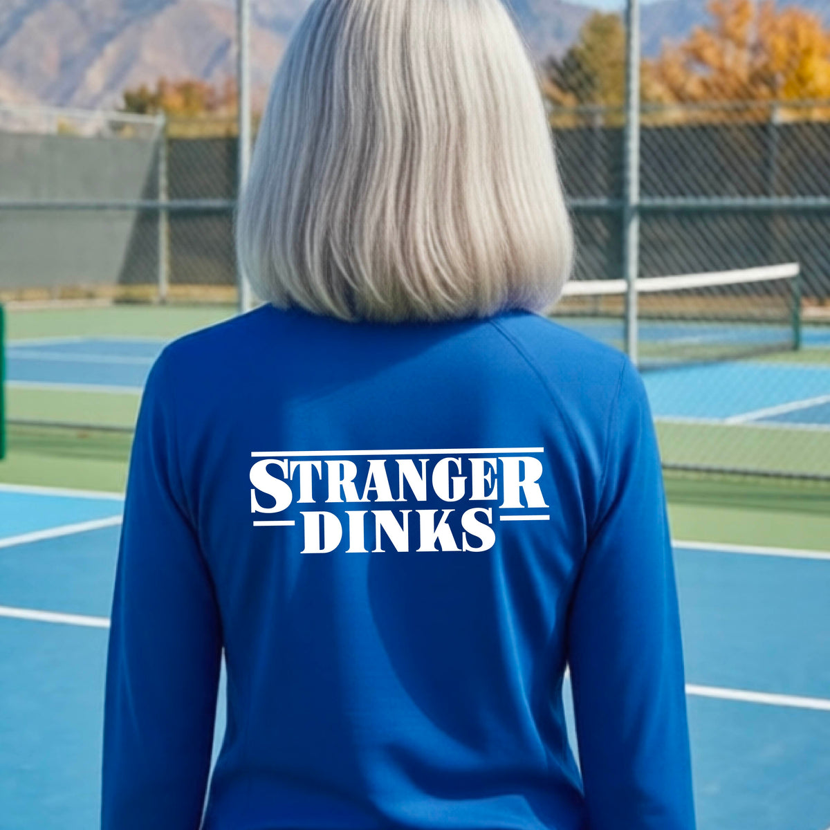 A person with short silver hair seen from behind wearing a royal blue quarter-zip long-sleeve shirt with a white graphic of the words “STRANGER DINKS” on the front of the shirt in the style of the popular TV Series Stranger Things. 