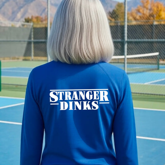 A person with short silver hair seen from behind wearing a royal blue quarter-zip long-sleeve shirt with a white graphic of the words “STRANGER DINKS” on the front of the shirt in the style of the popular TV Series Stranger Things. 