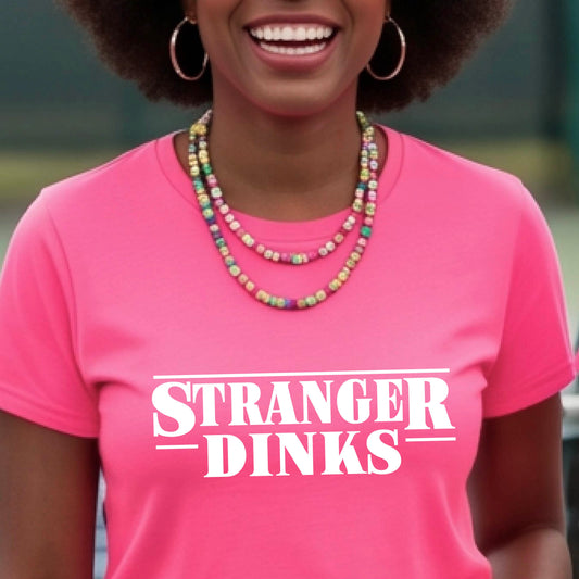 A woman on a pickleball court wearing a black short-sleeve t-shirt with a white graphic of the words “STRANGER DINKS” on the front of the shirt in the style of the popular TV Series Stranger Things. 