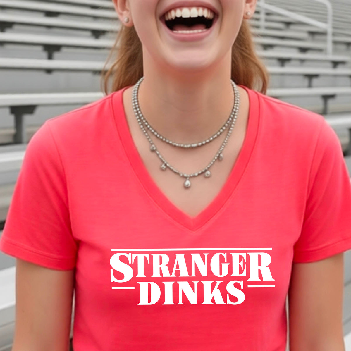 A woman wearing a coral pink short-sleeve t-shirt with a white graphic of the words “STRANGER DINKS” on the front of the shirt in the style of the popular TV Series Stranger Things. 