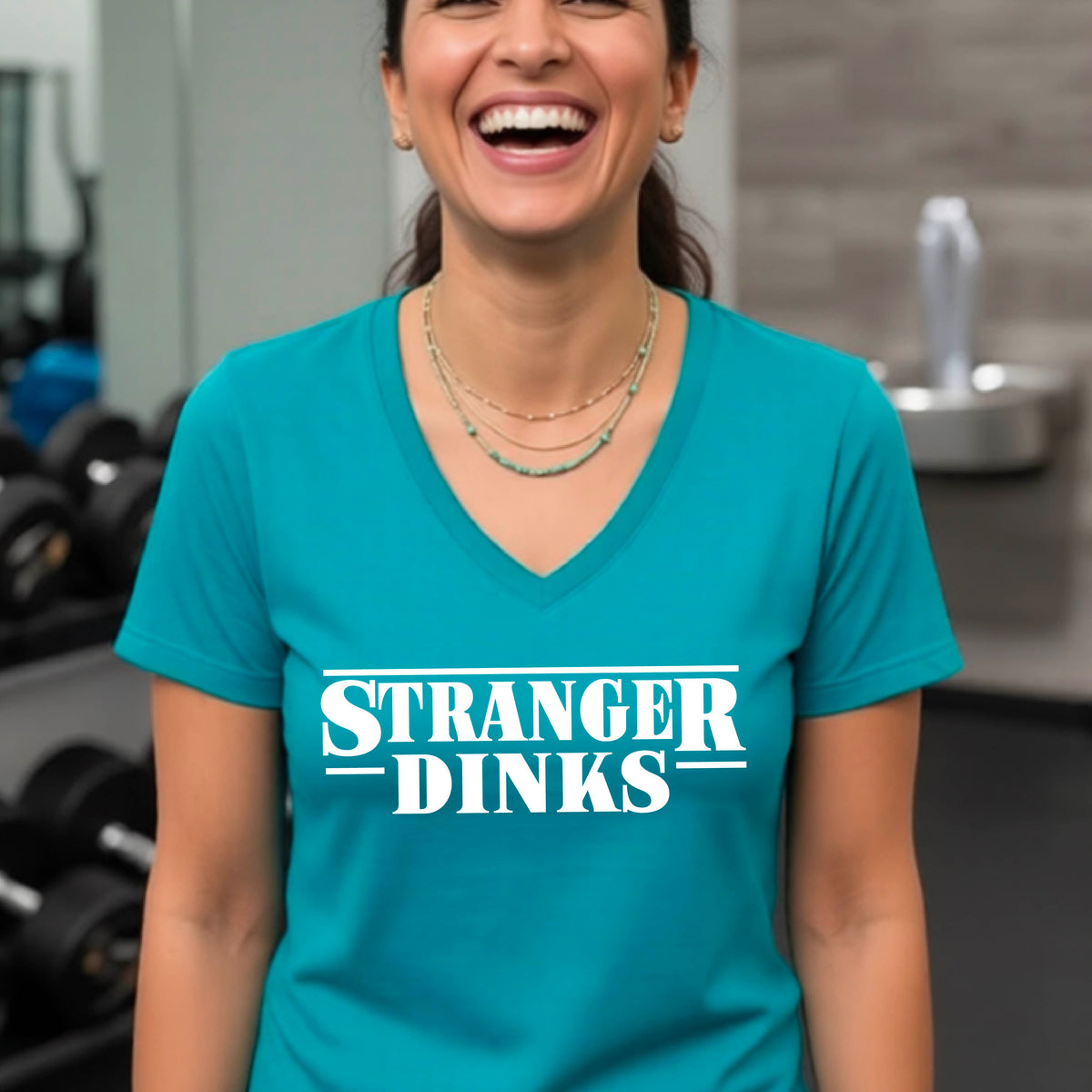 A woman wearing a teal short-sleeve t-shirt with a white graphic of the words “STRANGER DINKS” on the front of the shirt in the style of the popular TV Series Stranger Things. 