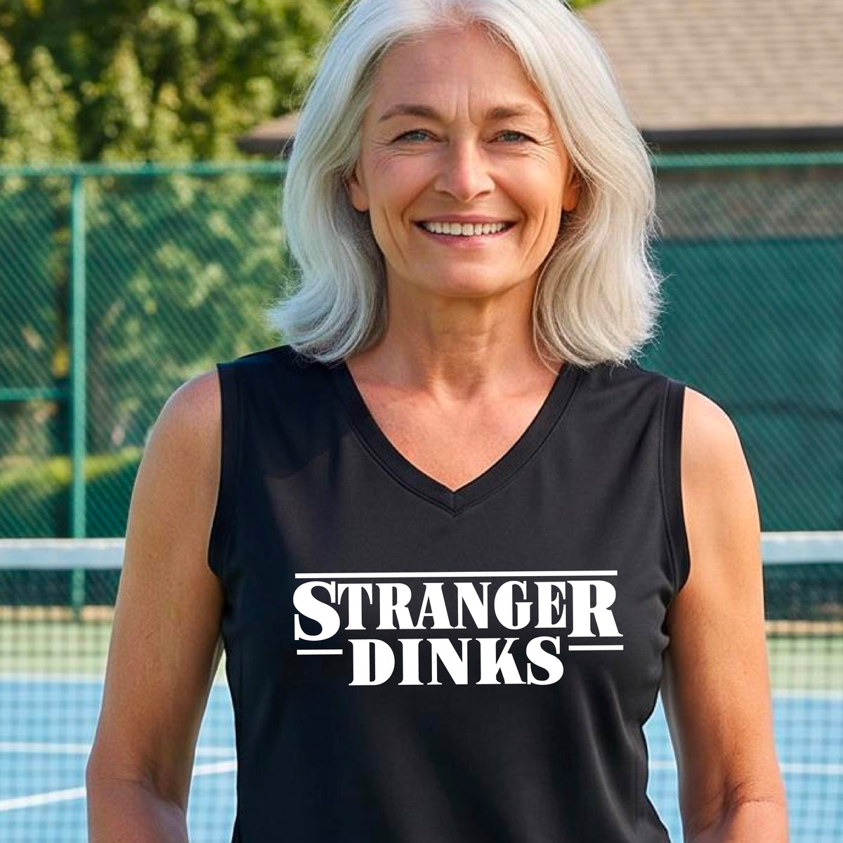 A woman on a pickleball court wearing a black sleeveless t-shirt with a white graphic of the words “STRANGER DINKS” on the front of the shirt in the style of the popular TV Series Stranger Things. 