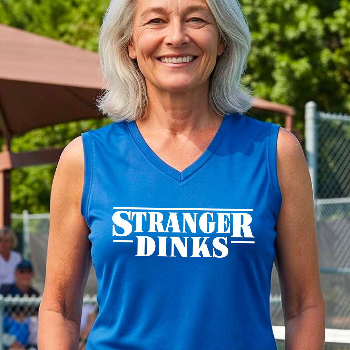 A woman on a pickleball court wearing a royal blue sleeveless t-shirt with a white graphic of the words “STRANGER DINKS” on the front of the shirt in the style of the popular TV Series Stranger Things. 