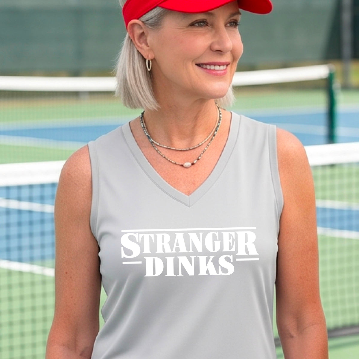 A woman on a pickleball court wearing a silver gray sleeveless t-shirt with a white graphic of the words “STRANGER DINKS” on the front of the shirt in the style of the popular TV Series Stranger Things. 