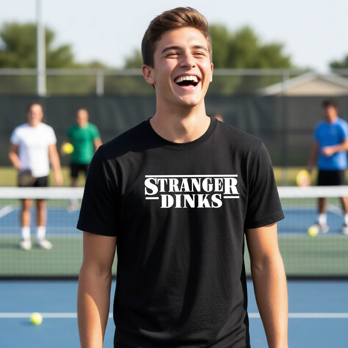 A young person on a pickleball court wearing a black short-sleeve t-shirt with a white graphic of the words “STRANGER DINKS” on the front of the shirt in the style of the popular TV Series Stranger Things. 