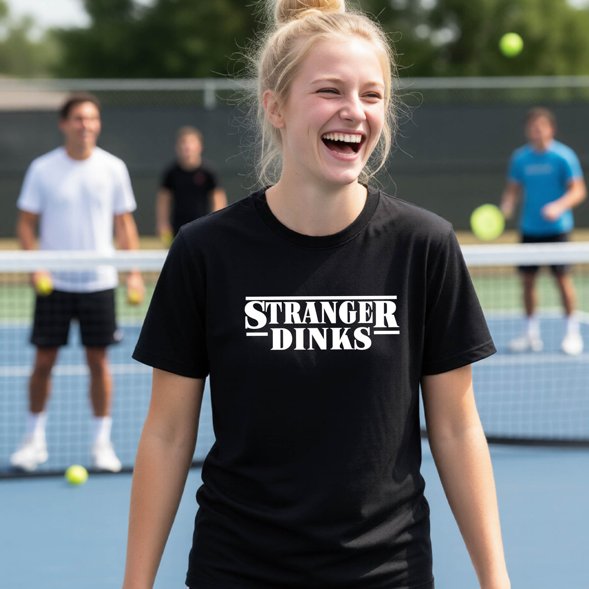 A young person on a pickleball court wearing a black short-sleeve t-shirt with a white graphic of the words “STRANGER DINKS” on the front of the shirt in the style of the popular TV Series Stranger Things. 