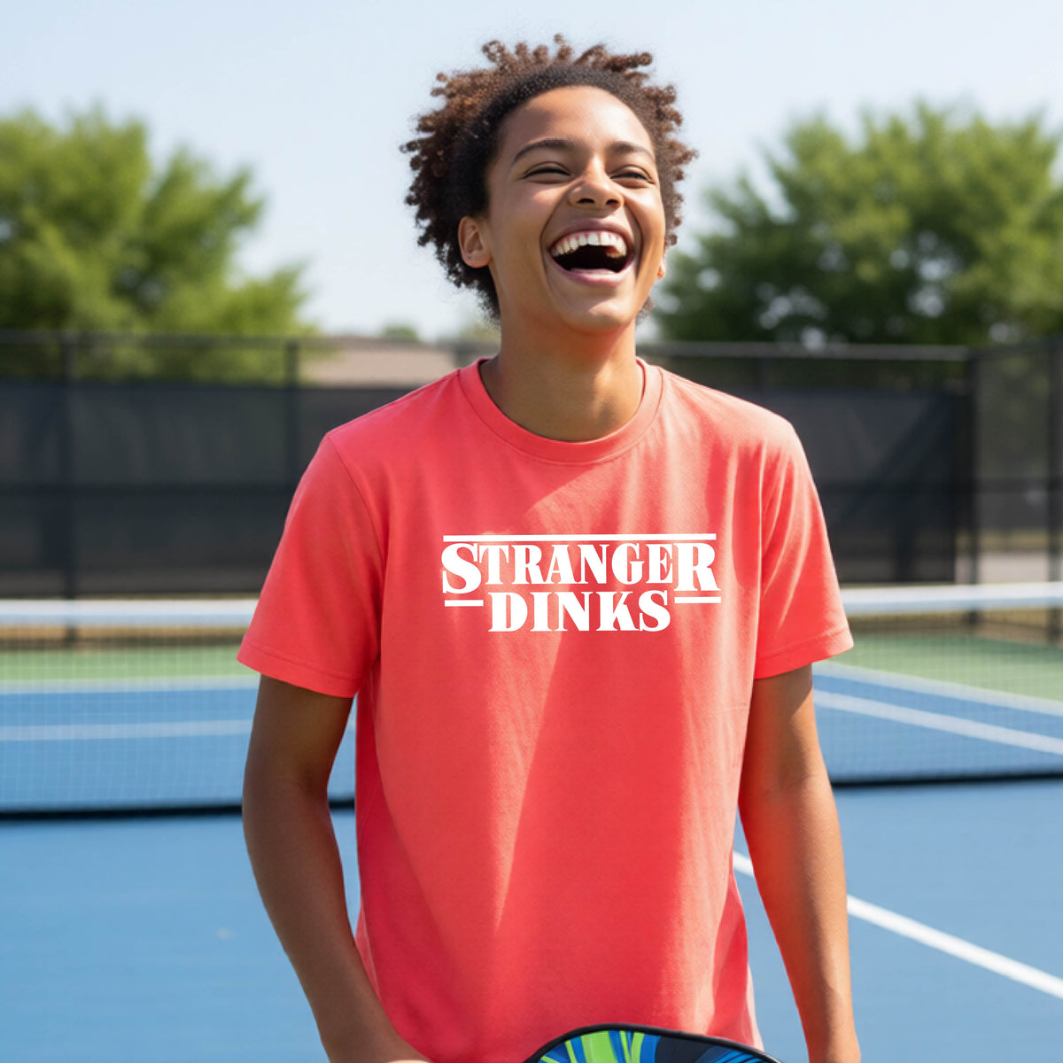 A young person on a pickleball court wearing a coral pink short-sleeve t-shirt with a white graphic of the words “STRANGER DINKS” on the front of the shirt in the style of the popular TV Series Stranger Things. 