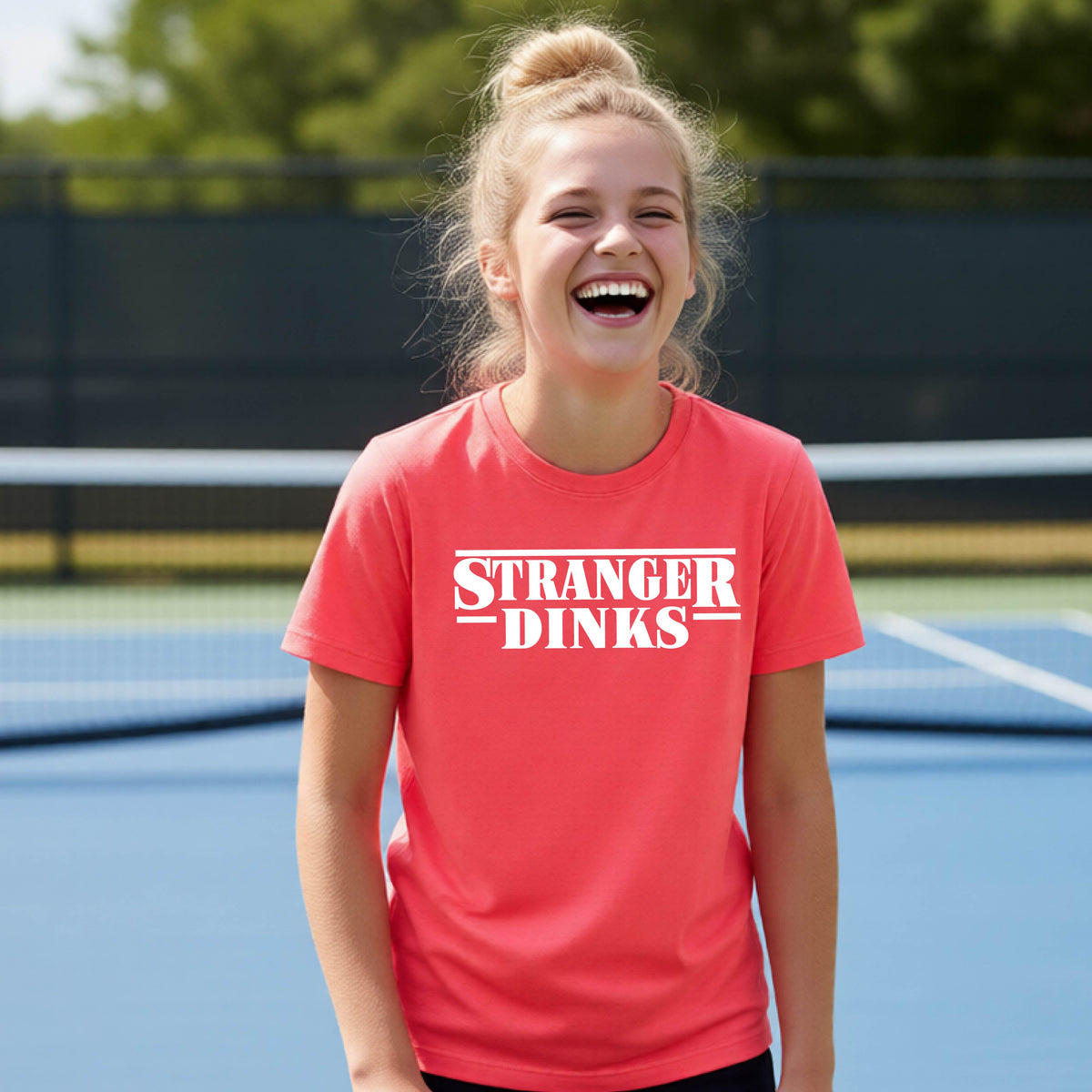 A young person on a pickleball court wearing a coral pink short-sleeve t-shirt with a white graphic of the words “STRANGER DINKS” on the front of the shirt in the style of the popular TV Series Stranger Things. 