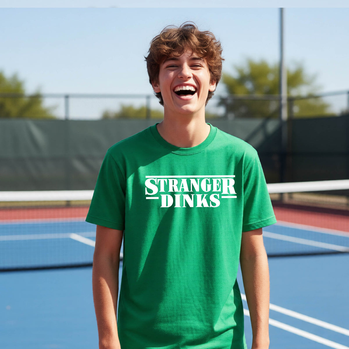 A young person on a pickleball court wearing a green short-sleeve t-shirt with a white graphic of the words “STRANGER DINKS” on the front of the shirt in the style of the popular TV Series Stranger Things. 