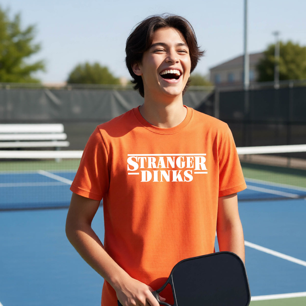 A young person on a pickleball court wearing an orange short-sleeve t-shirt with a white graphic of the words “STRANGER DINKS” on the front of the shirt in the style of the popular TV Series Stranger Things. 