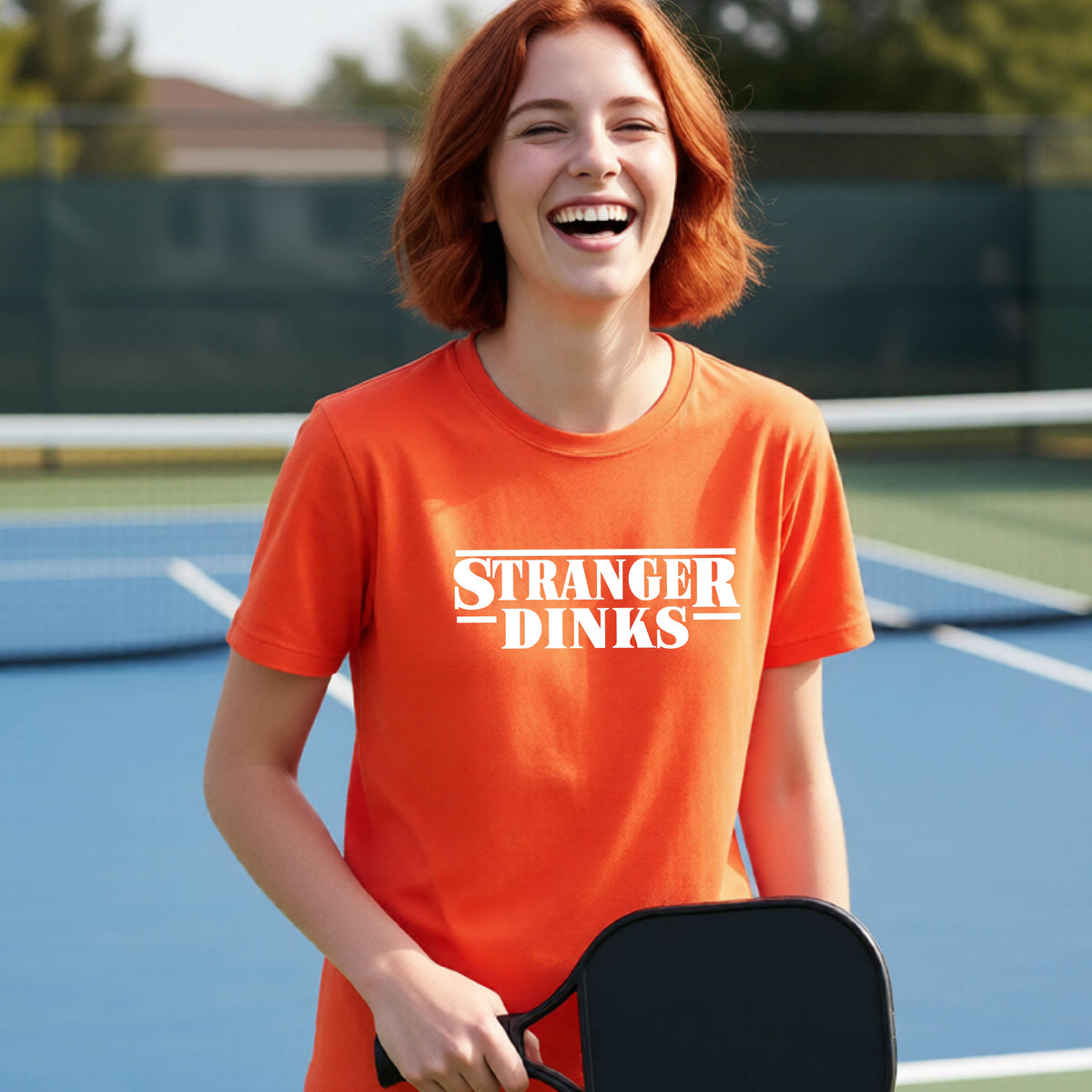 A young person on a pickleball court wearing an orange short-sleeve t-shirt with a white graphic of the words “STRANGER DINKS” on the front of the shirt in the style of the popular TV Series Stranger Things. 