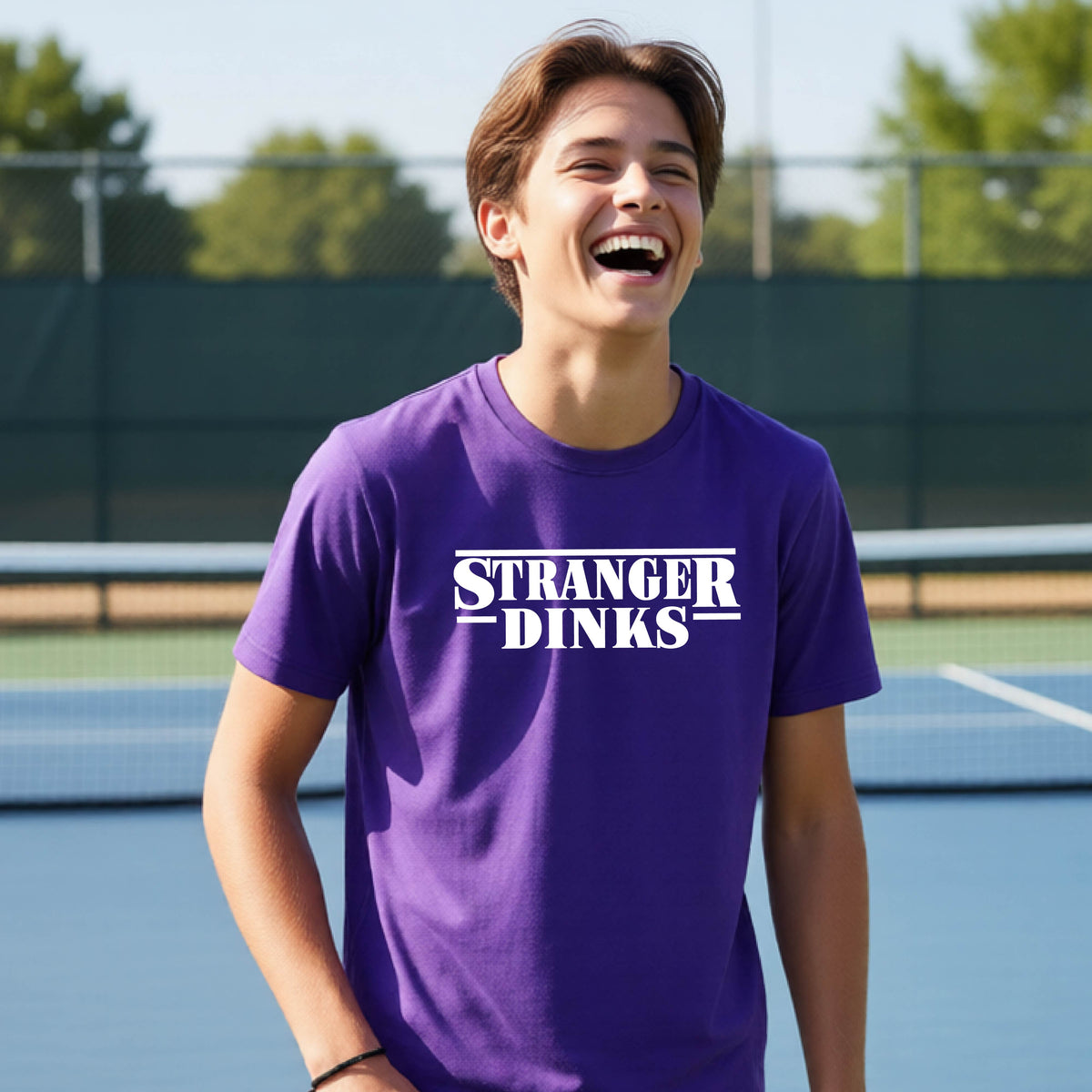 A young person on a pickleball court wearing a purple short-sleeve t-shirt with a white graphic of the words “STRANGER DINKS” on the front of the shirt in the style of the popular TV Series Stranger Things. 