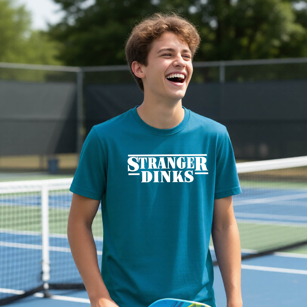 A young person on a pickleball court wearing a teal short-sleeve t-shirt with a white graphic of the words “STRANGER DINKS” on the front of the shirt in the style of the popular TV Series Stranger Things. 