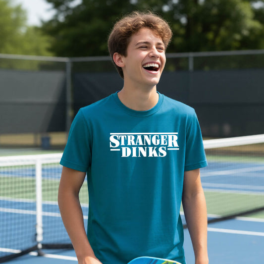 A young person on a pickleball court wearing a teal short-sleeve t-shirt with a white graphic of the words “STRANGER DINKS” on the front of the shirt in the style of the popular TV Series Stranger Things. 